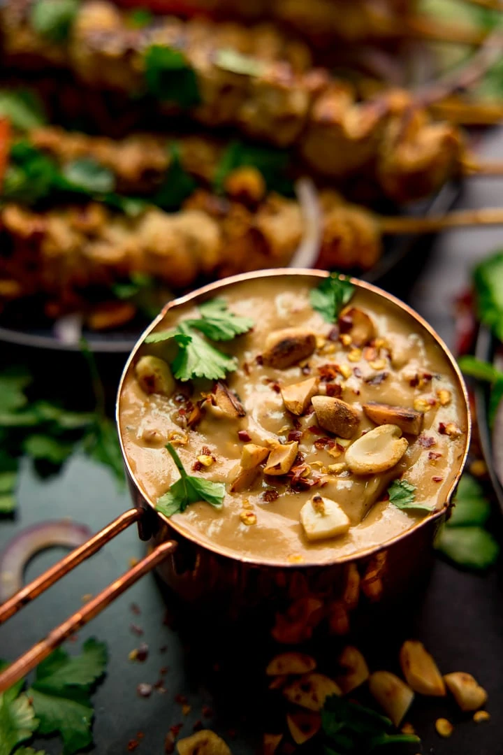 Satay sauce in a small copper pan topped with peanuts and coriander. Chicken skewers in the background