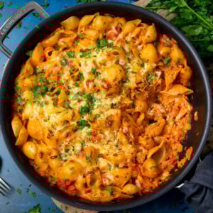 A frying pan filled with Creamy Tomato Tuna Pasta Bake—pasta shells in rich tomato sauce with tuna, topped with melted cheese and chopped parsley.