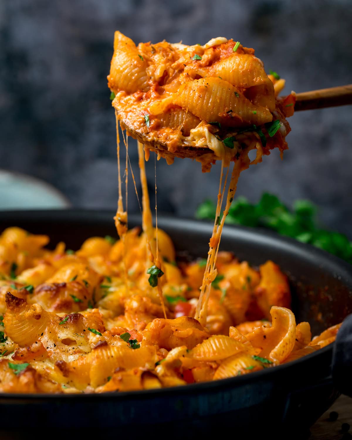 A pan of Creamy Tomato Tuna Pasta Bake with melted cheese, served in tomato sauce, is being served with a wooden spoon. The cheese is stretching as the portion is being lifted from the pan.