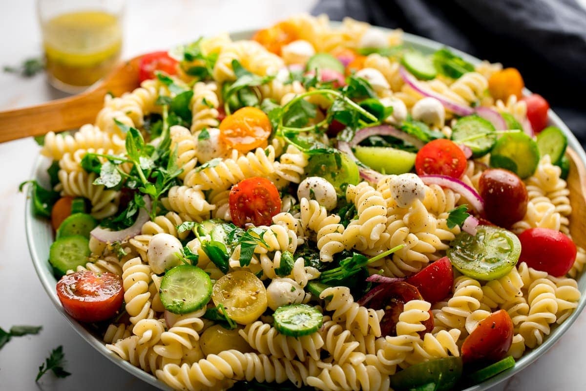 wide image of a bowl of pasta salad with tomatoes, mozzarella balls, pea shoots and cucumber.