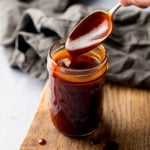 Spoonful being taken from a jar of homemade bbq sauce, sitting on a wooden board