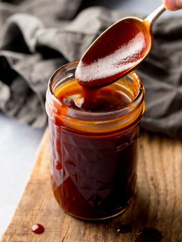 Spoonful being taken from a jar of homemade bbq sauce, sitting on a wooden board