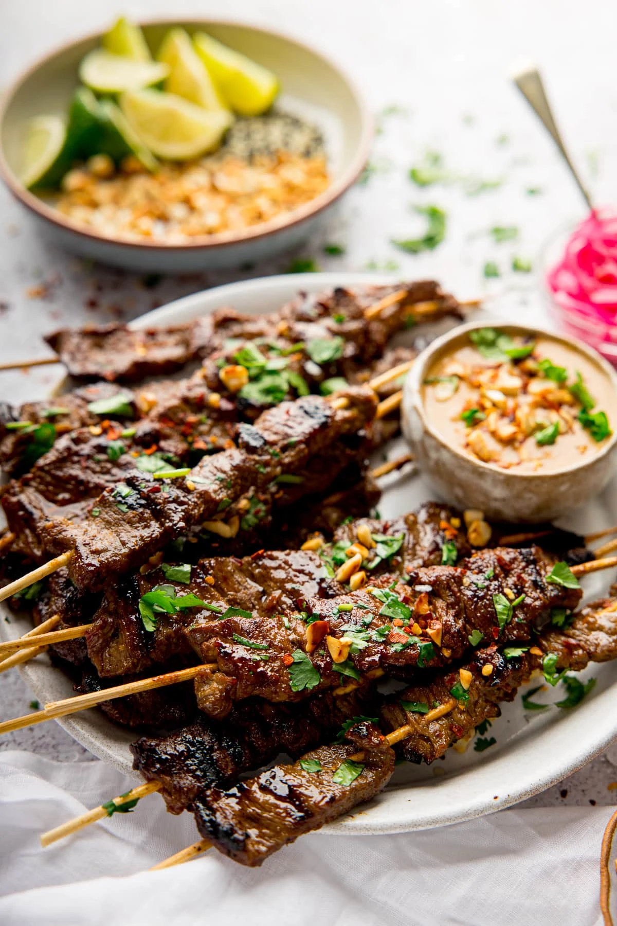 beef satay on a white plate with satay sauce. Bowl of chopped peanuts and lime in background.