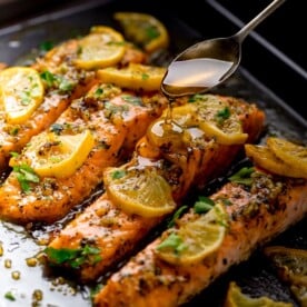 Baked salmon on a tray topped with lemon slices. Honey being drizzled on top with a spoon.