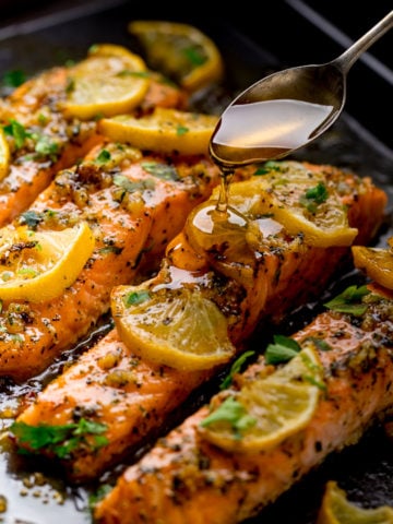 Baked salmon on a tray topped with lemon slices. Honey being drizzled on top with a spoon.