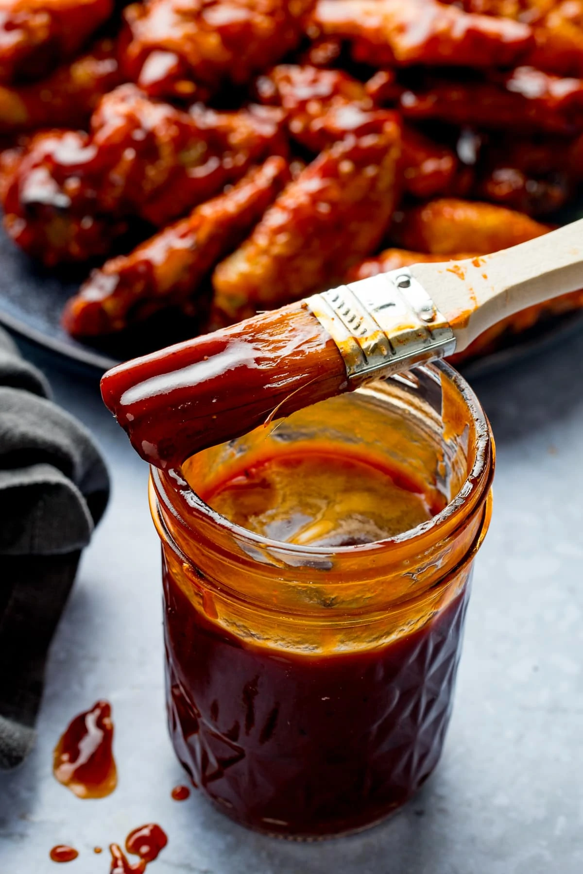 Jar of homemade bbq sauce with a basting brush on top, chicken wings in the background