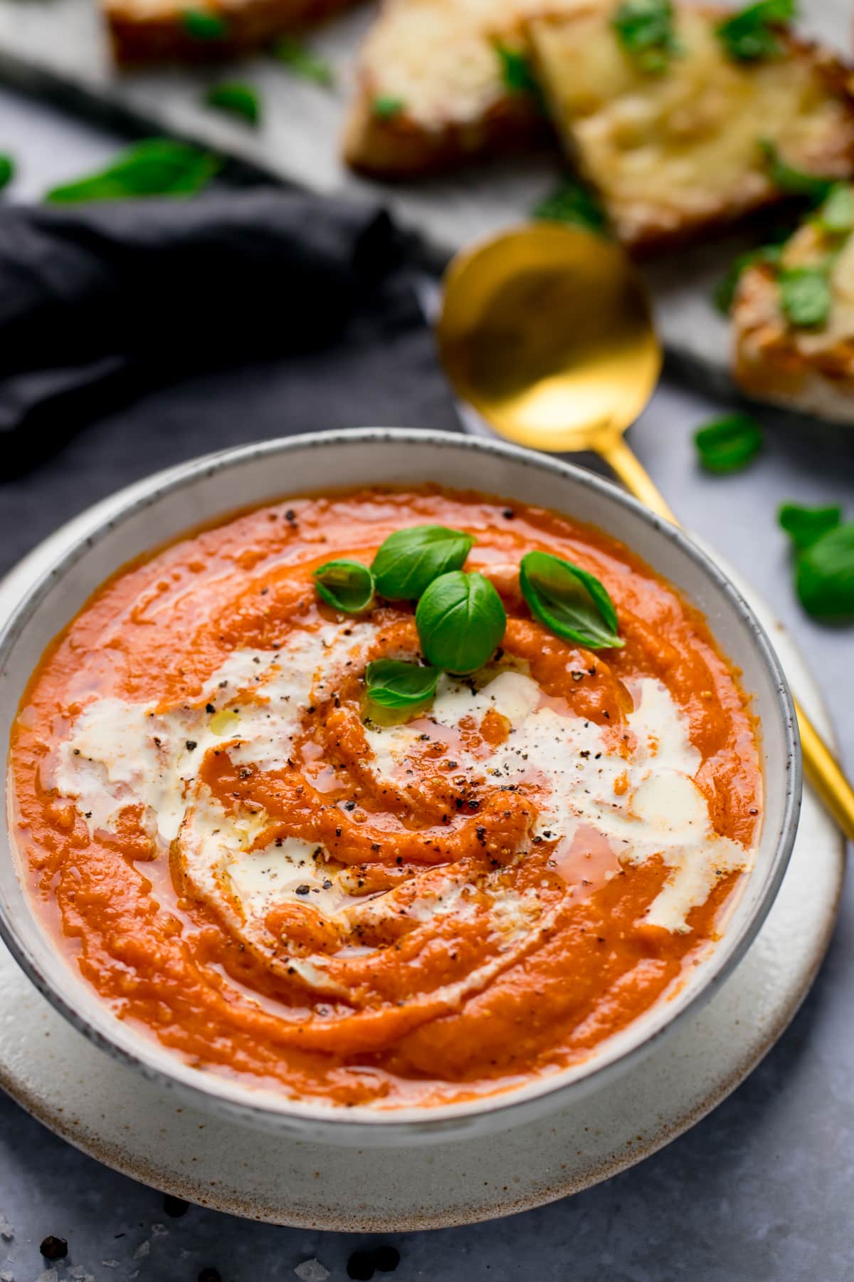 Lush bowl of wholesome tomato soup covered with rich cream and basil leaves with a golden spoon. A slate of grilled cheese toast covered with basil leaves in the background.