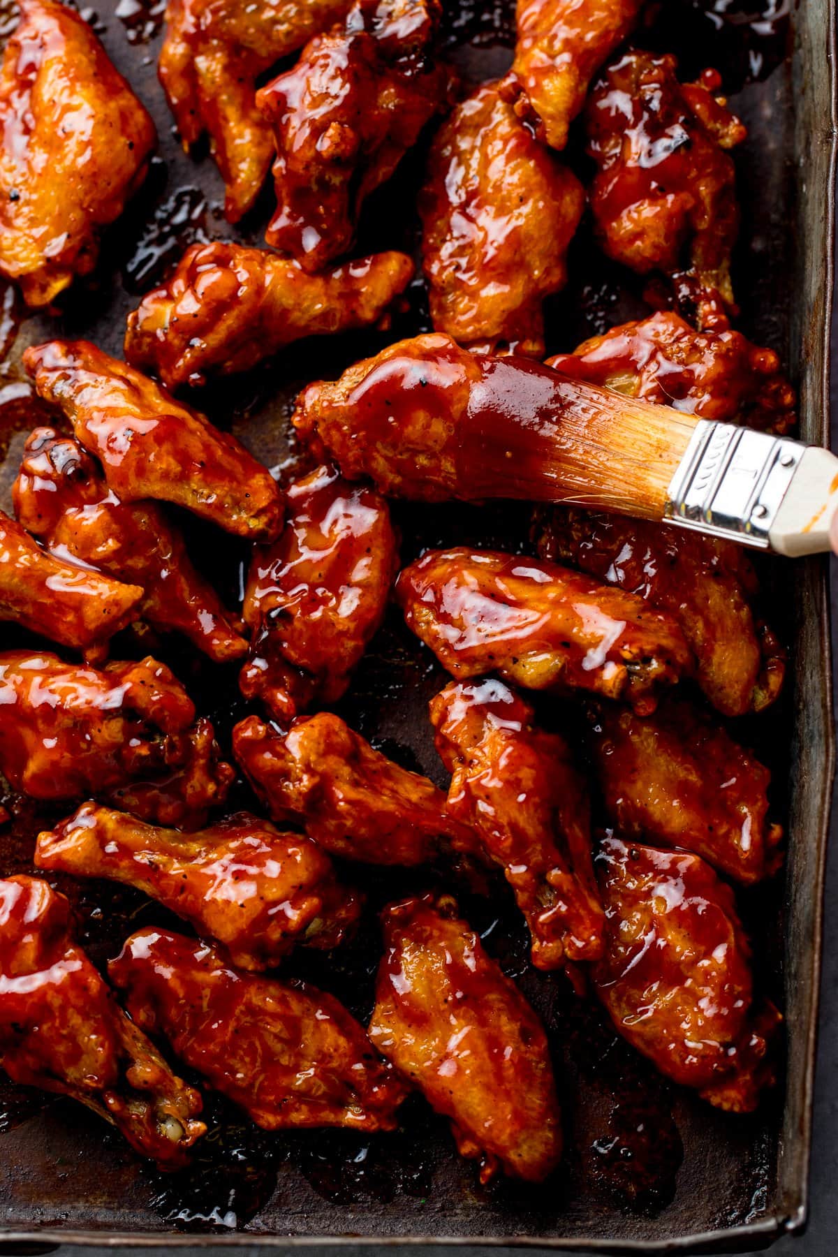 Tall image of BBQ chicken wings on a baking tray with sauce being brushed on