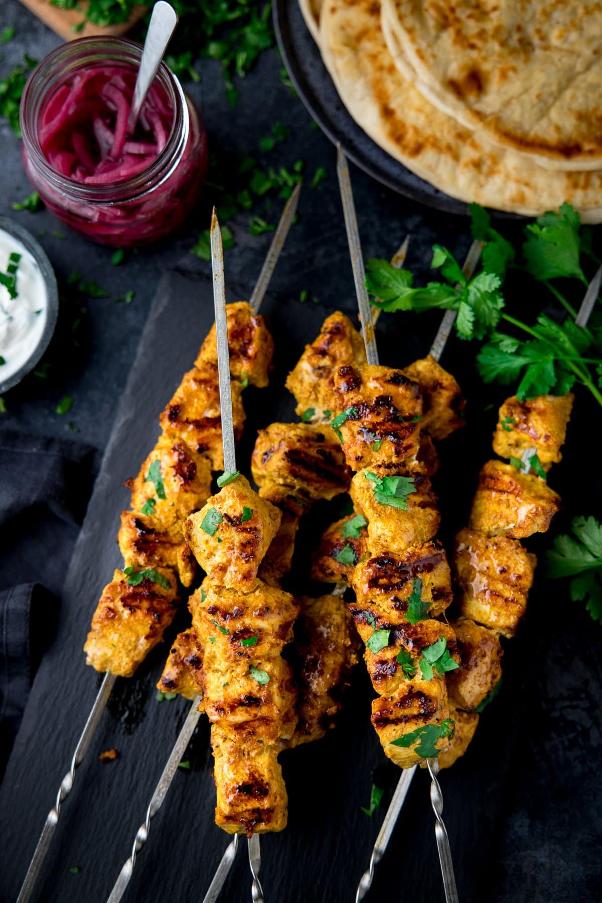 chicken tikka skewers on a dark background with pita bread and a jar of pickled red onion slices