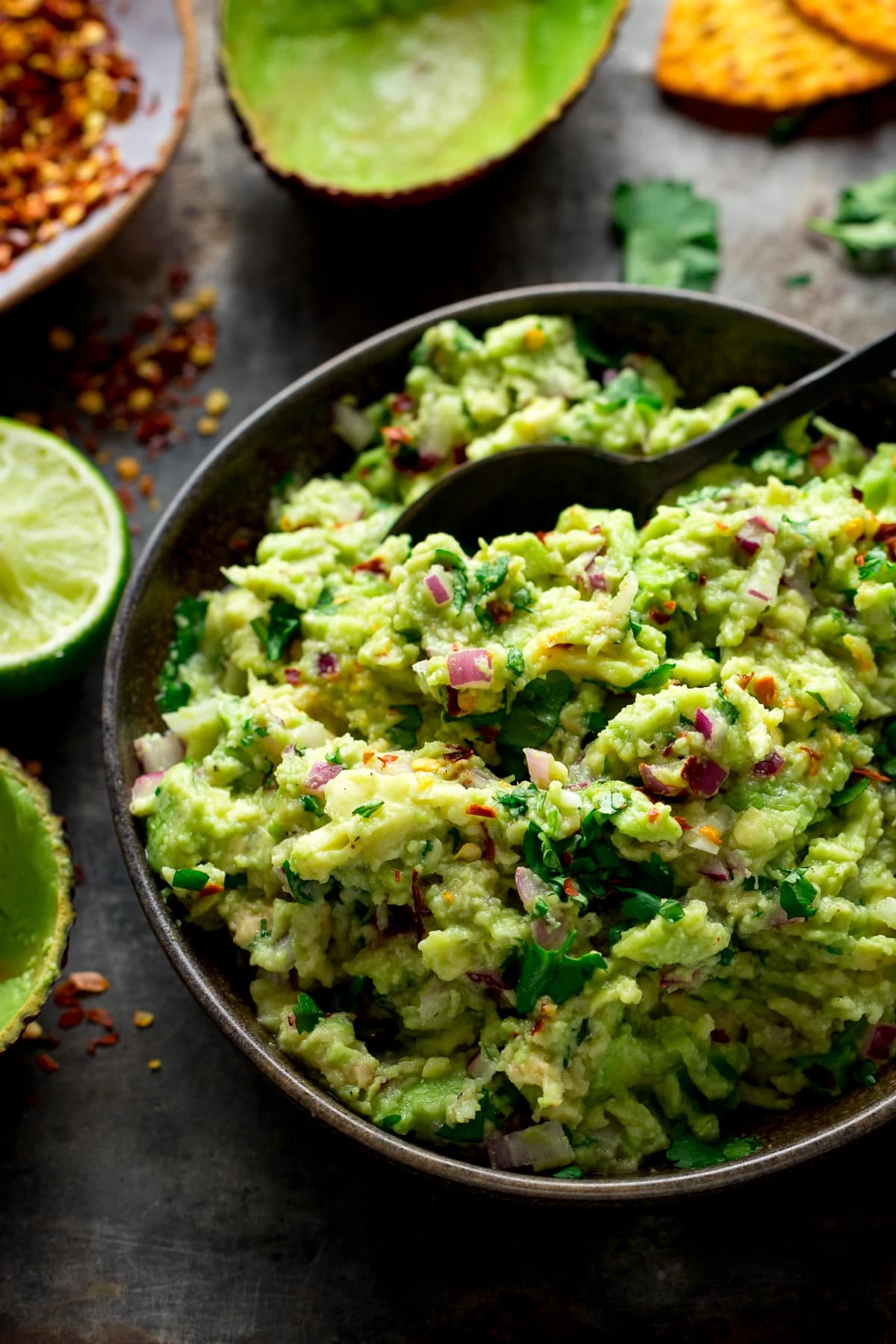 Guacamole in a brown bowl with a spoon, and avocadao shells and limes at the side.