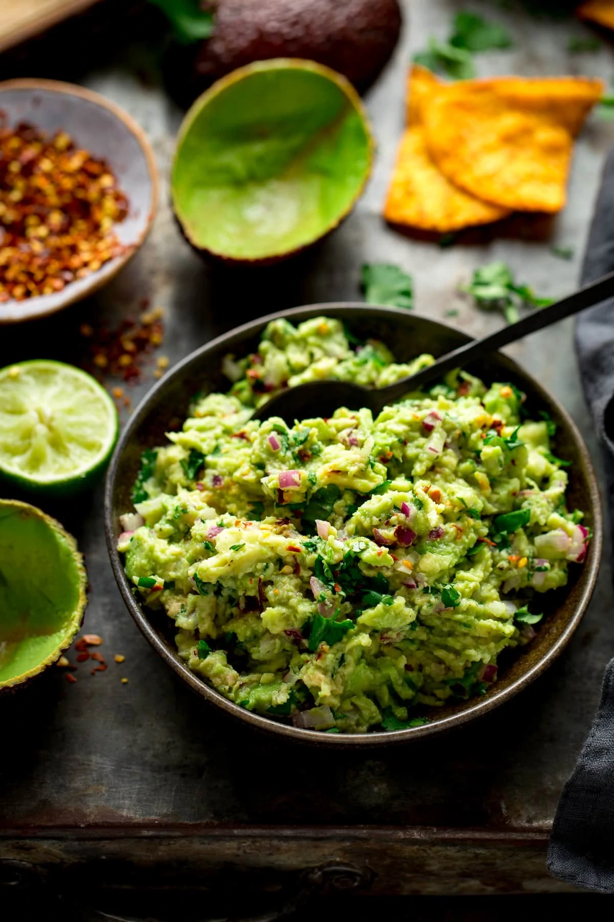 Guacamole in a brown bowl with a spoon on a dark table with limes and avocado shells next to the bowl