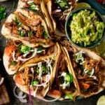 Square image of a plate of Lamb Barbacoa Carnita Tacos with a bowl of guacamole