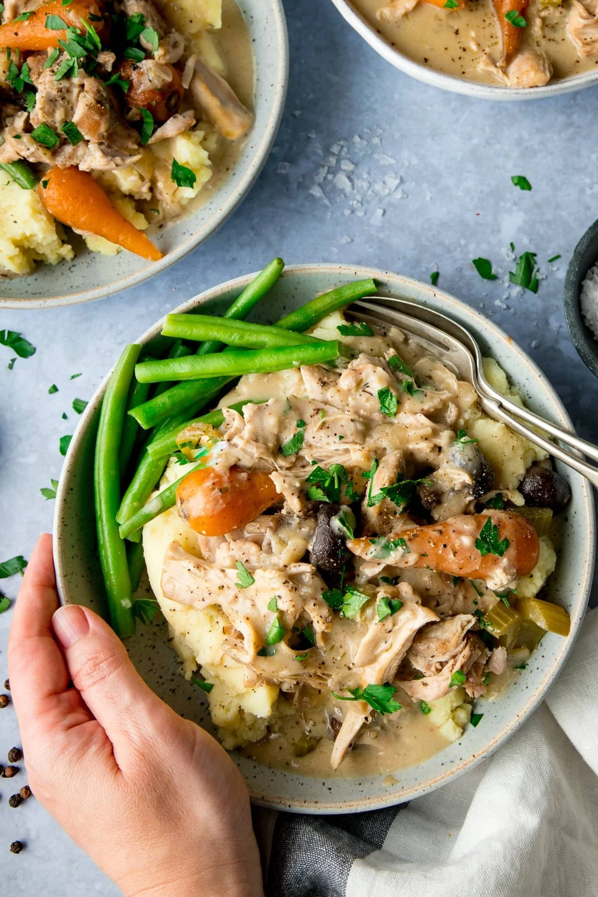 Hand holding a bowl of chicken casserole with mashed potatoes and green beans on a light blue background