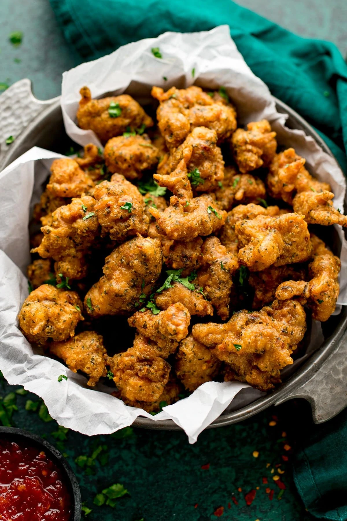 Bowl of chicken pakora with a green napkin on a green background. Chilli sauce next to the bowl.