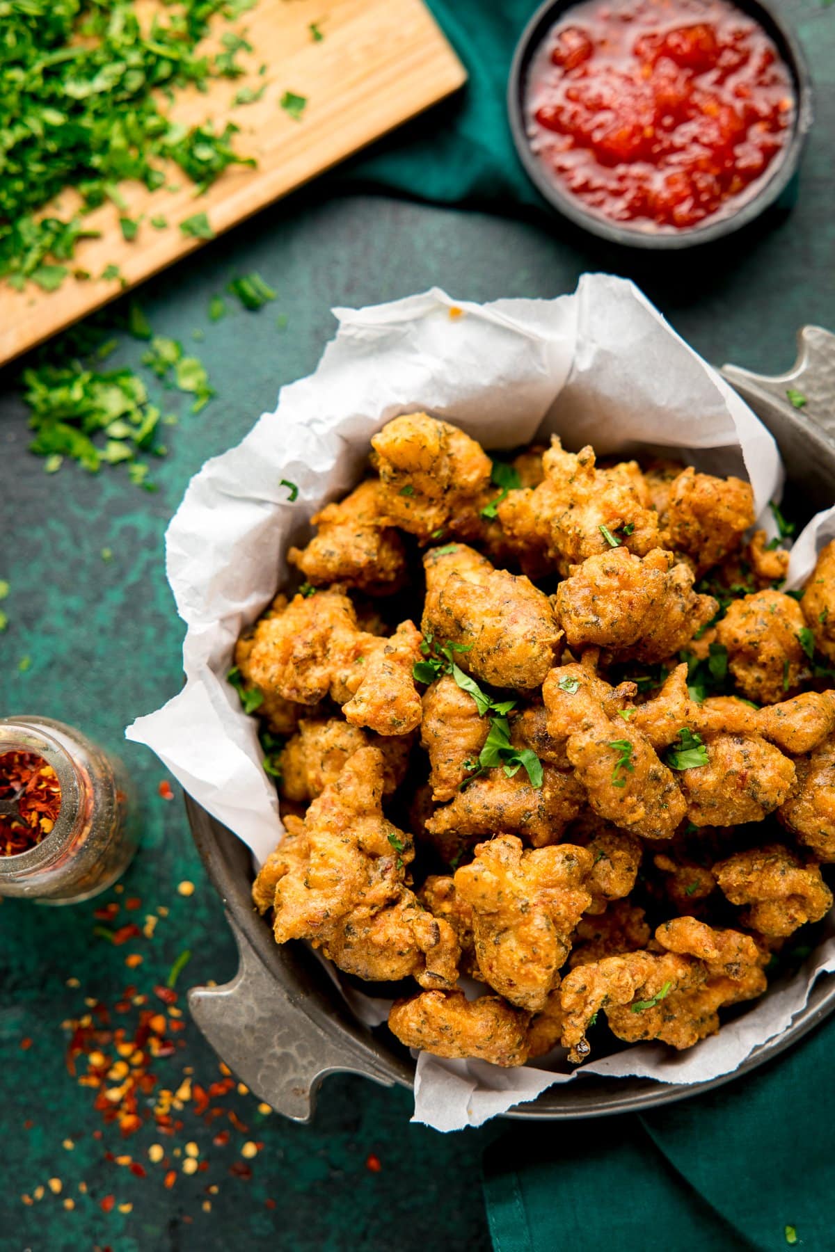 Overhead image of a bowl of chicken pakora on a green background with chilli sauce, chilli flakes and coriander scattered around
