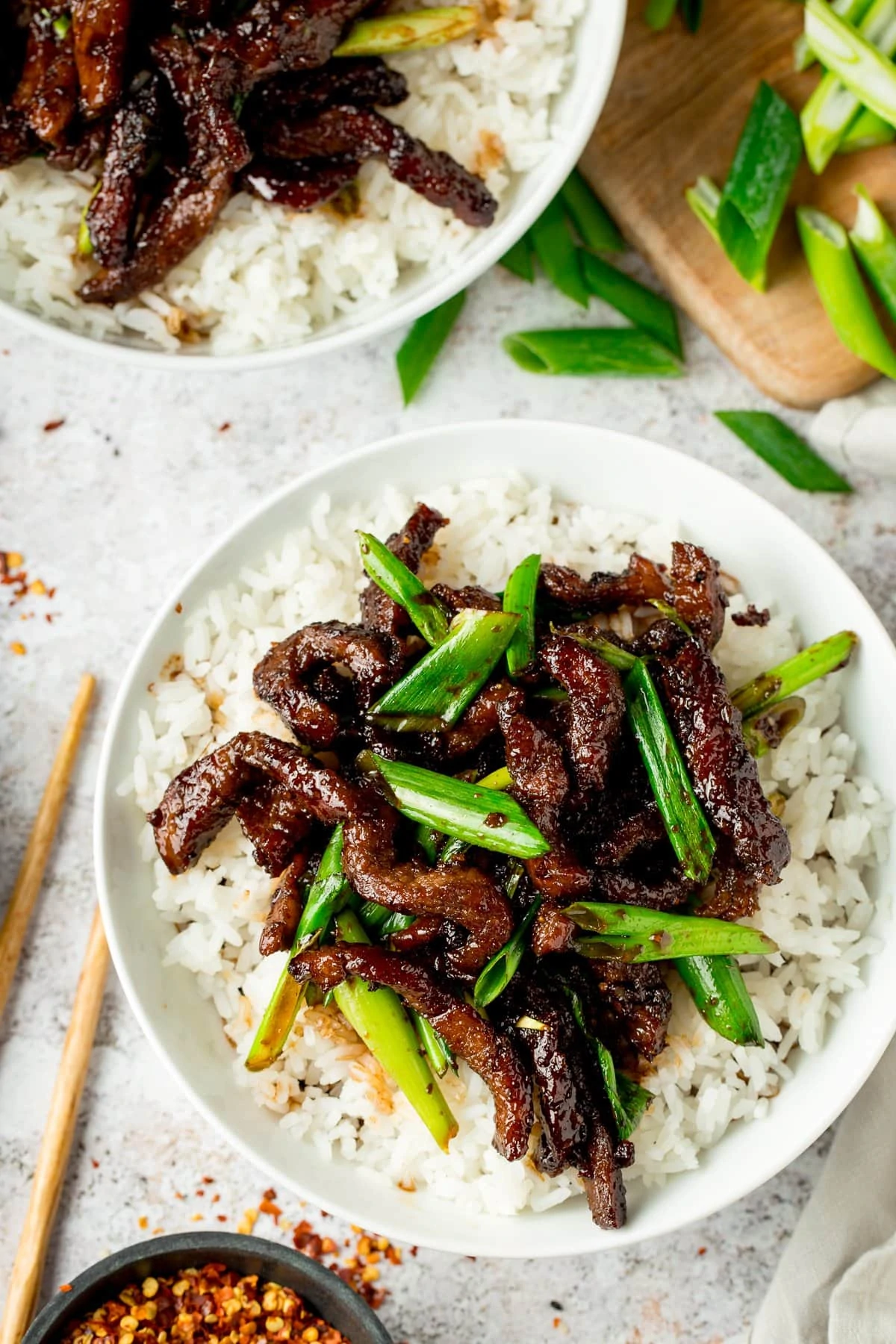 Mongolian Beef and rice in a white bowl on a light background with chopsticks and ingredients scattered around