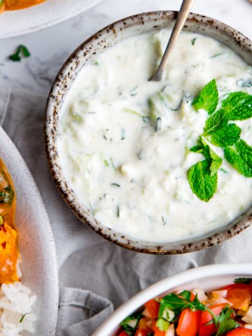 Raita in a grey bowl with mint leaves on top and bowls of food around the raita