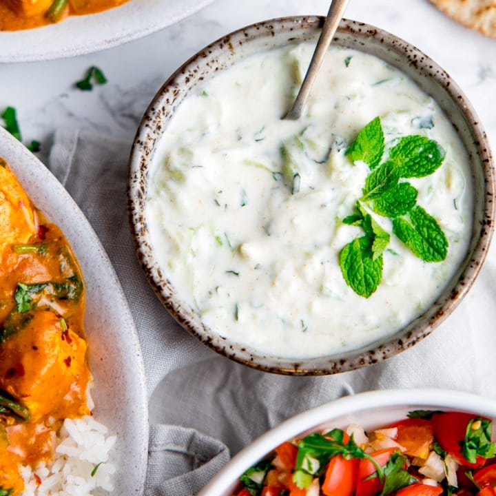 Raita in a grey bowl with mint leaves on top and bowls of food around the raita