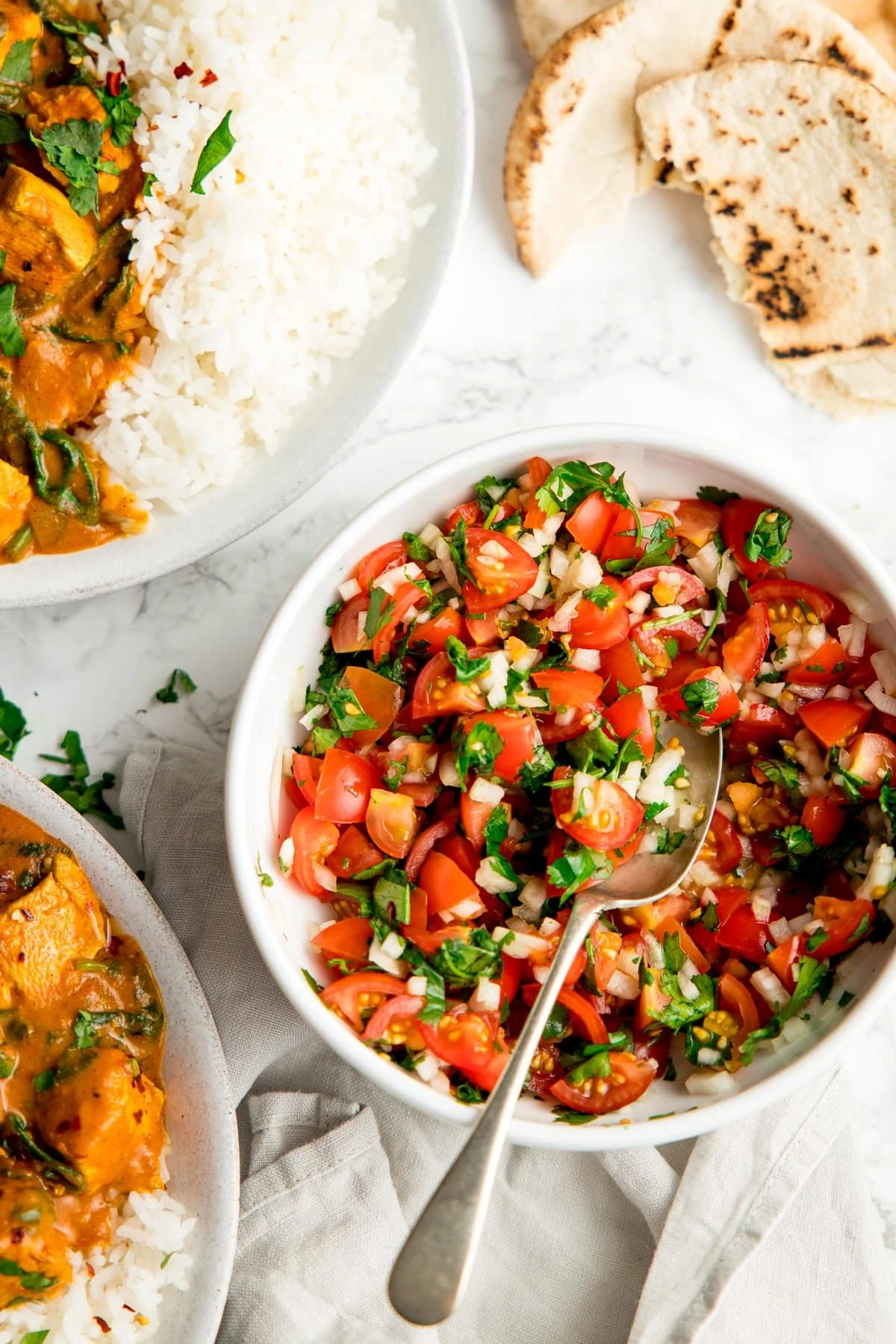 tomato, onion and coriander chopped salad in a white bowl with a spoon on a white background. Pita and curry dishes a little bit in shot