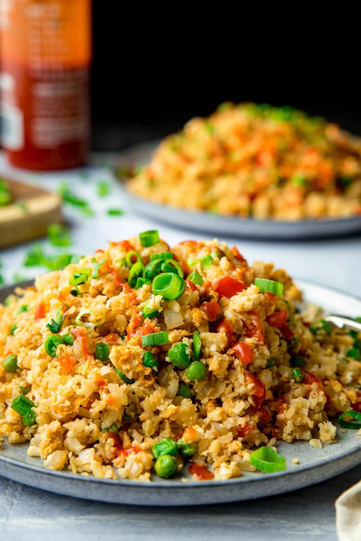 Cauliflower fried rice piled up on a plate with a further plate and chilli sauce bottle in the background