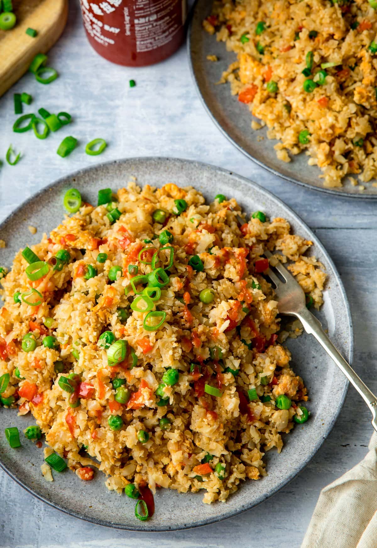 Cauliflower fried rice topped with spring onions on a light grey background. Further plate partially in shot.