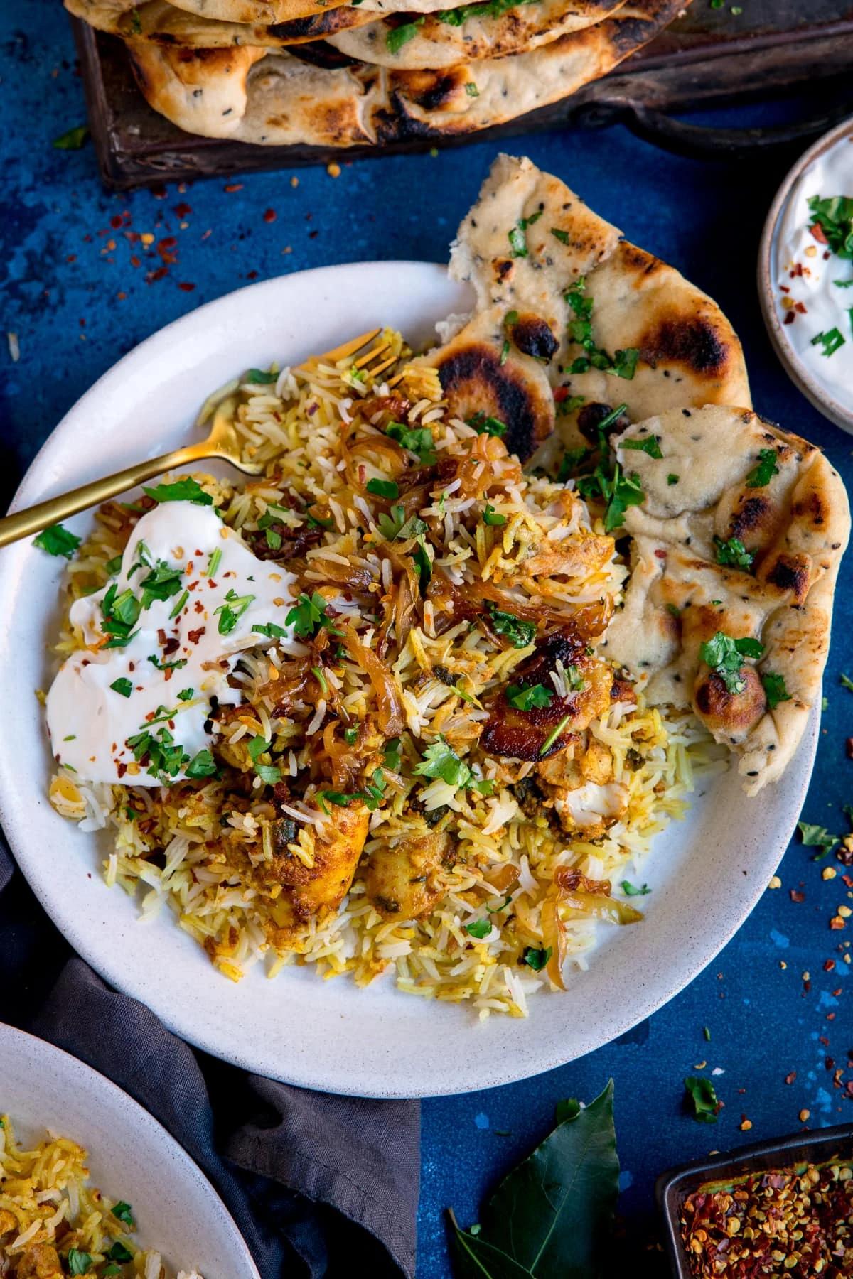 chicken biryani and natural yogurt on a plate with naan bread. Plate is on a blue background.