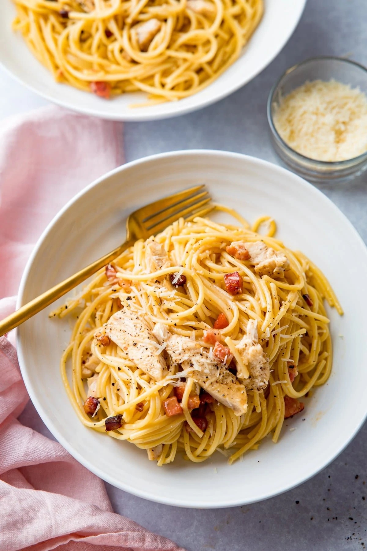 chicken carbonara in a bowl next to a pink napkin. Further bowl and a bowl of parmesan just in shot