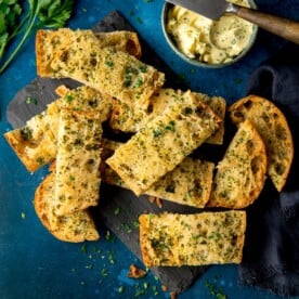 Sliced of garlic bread on a dark blue background with a bowl of garlic butter and knife at the top of the frame