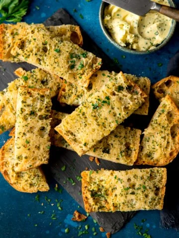 Sliced of garlic bread on a dark blue background with a bowl of garlic butter and knife at the top of the frame