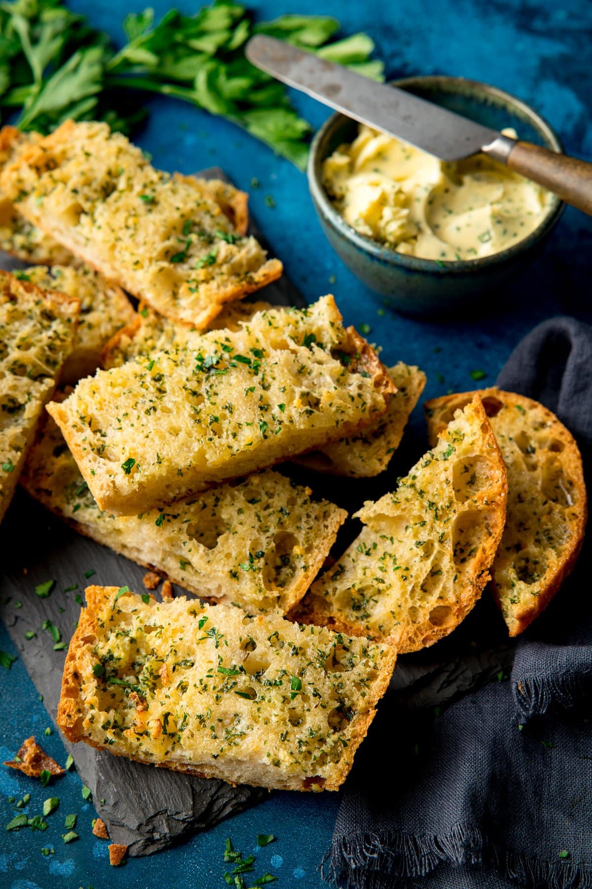 Slices of garlic bread on a slate board on a dark blue background with bowl of garlic butter and knife next to it