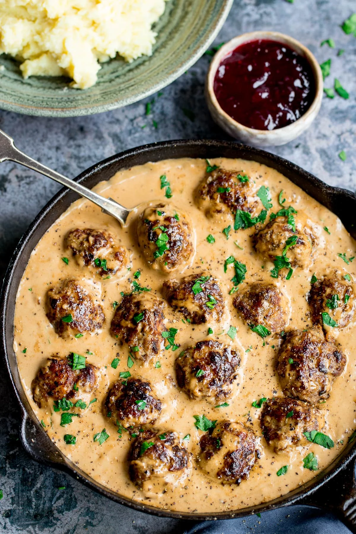 Swedish meatballs and gravy in a pan next to a pot of lingonberry jelly and mashed potatoes