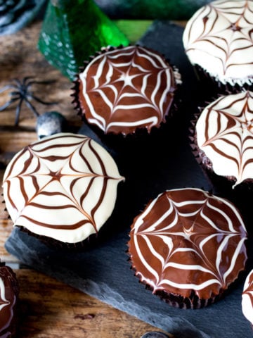 Chocolate Halloween cupcakes with spiderweb patterns in the topping placed on a dark slate background with toy skulls and spiders in the background.