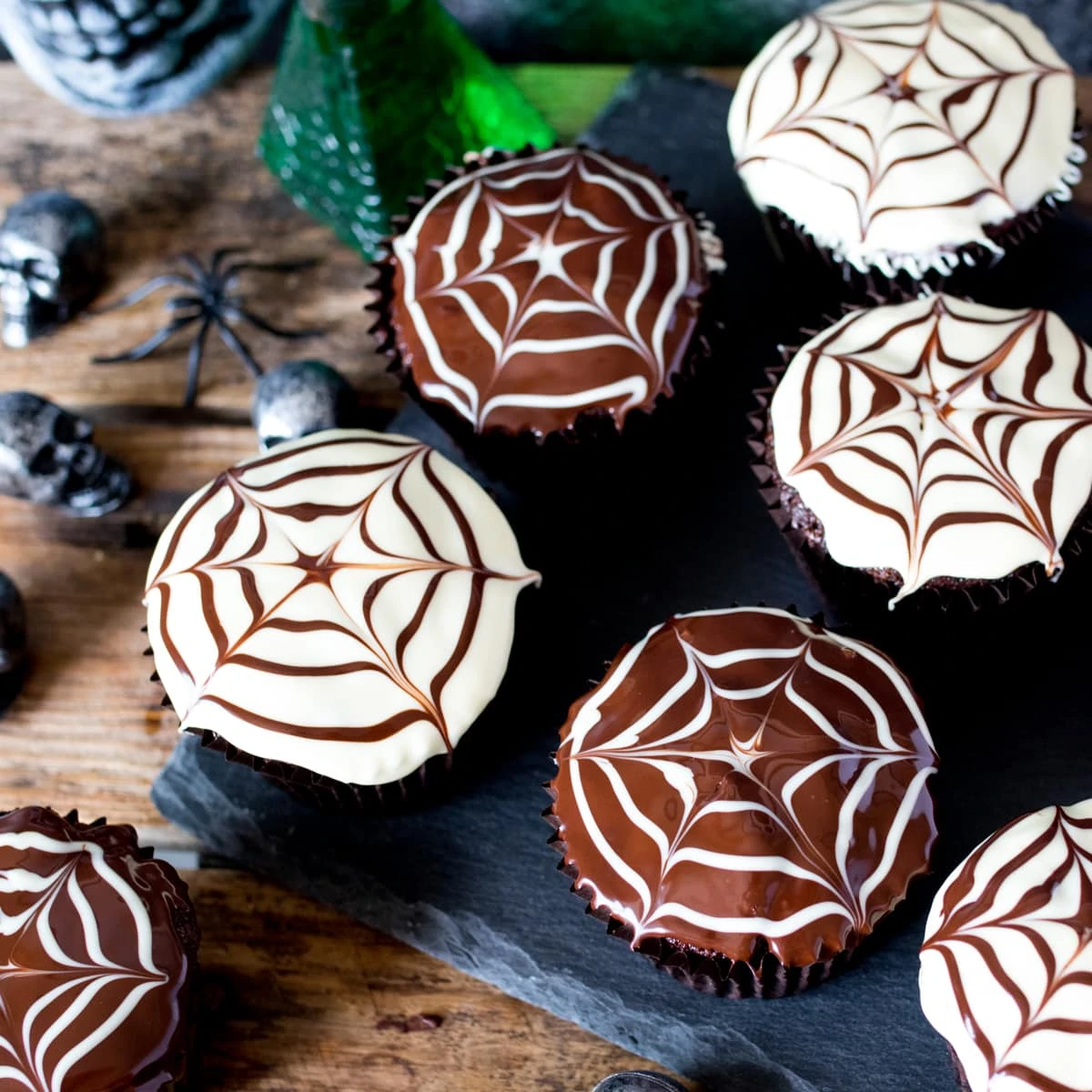 Chocolate Halloween cupcakes with spiderweb patterns in the topping placed on a dark slate background with toy skulls and spiders in the background.