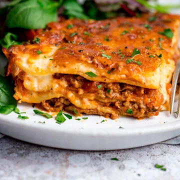 Portion of lasagne on a white plate with salad in the background