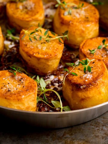 Square image of fondant potatoes in a frying pan, topped with fresh thyme and salt