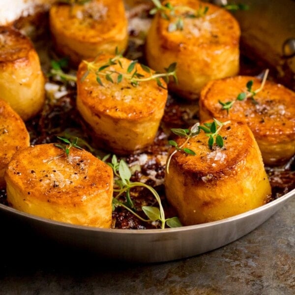 Square image of fondant potatoes in a frying pan, topped with fresh thyme and salt