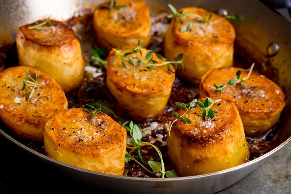 Wide image of fondant potatoes in a pan topped with fresh thyme