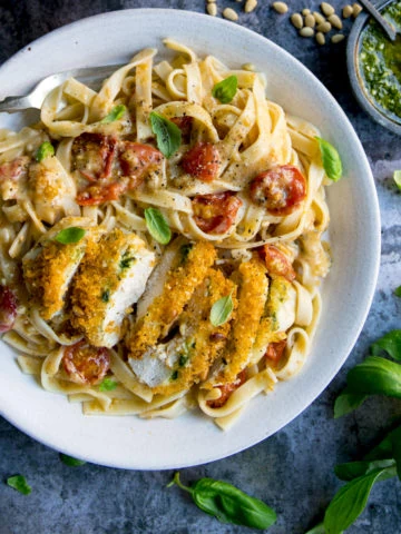 Crispy chicken, sliced and served in a creamy pesto tomato sauce on top of pasta on a white plate. There are ingredients scattered around. The plate is on a blue mottled background.