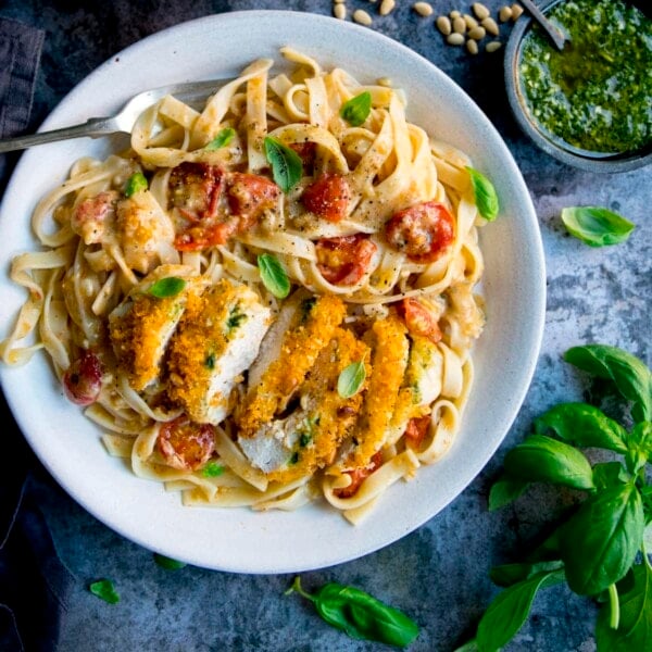 Crispy chicken, sliced and served in a creamy pesto tomato sauce on top of pasta on a white plate. There are ingredients scattered around. The plate is on a blue mottled background.