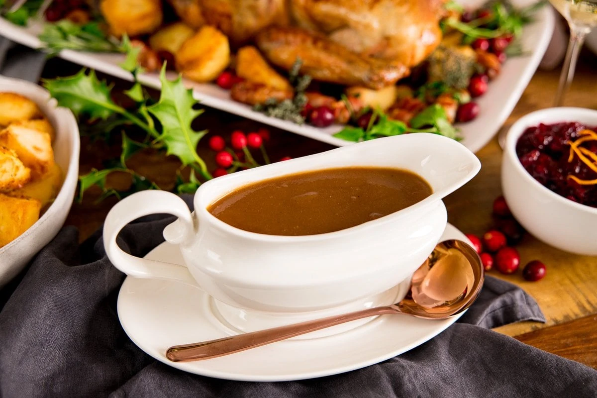 A white gravy boat filled with gravy on a table with a roast turkey and side dishes around the gravy boat. Copper spoon resting on the gravy boat saucer.