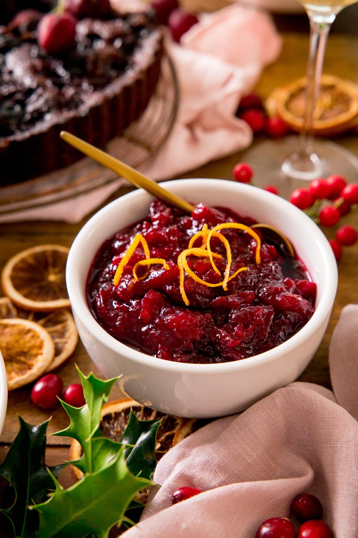 Cranberry sauce in a white bowl, topped with orange zest. Festive table decorations scattered around the bowl.
