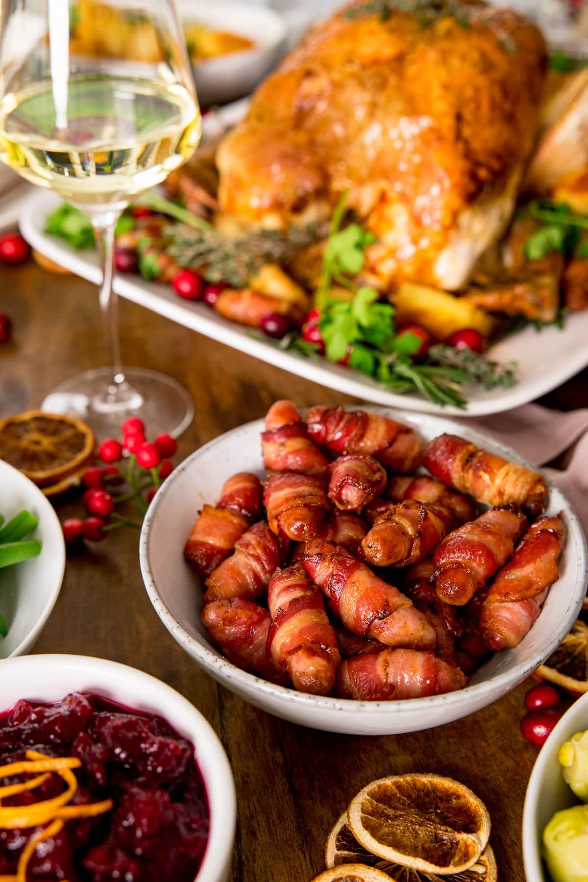 Sausages wrapped in bacon in a white bowl on a table with wine, turkey and cranberry sauce
