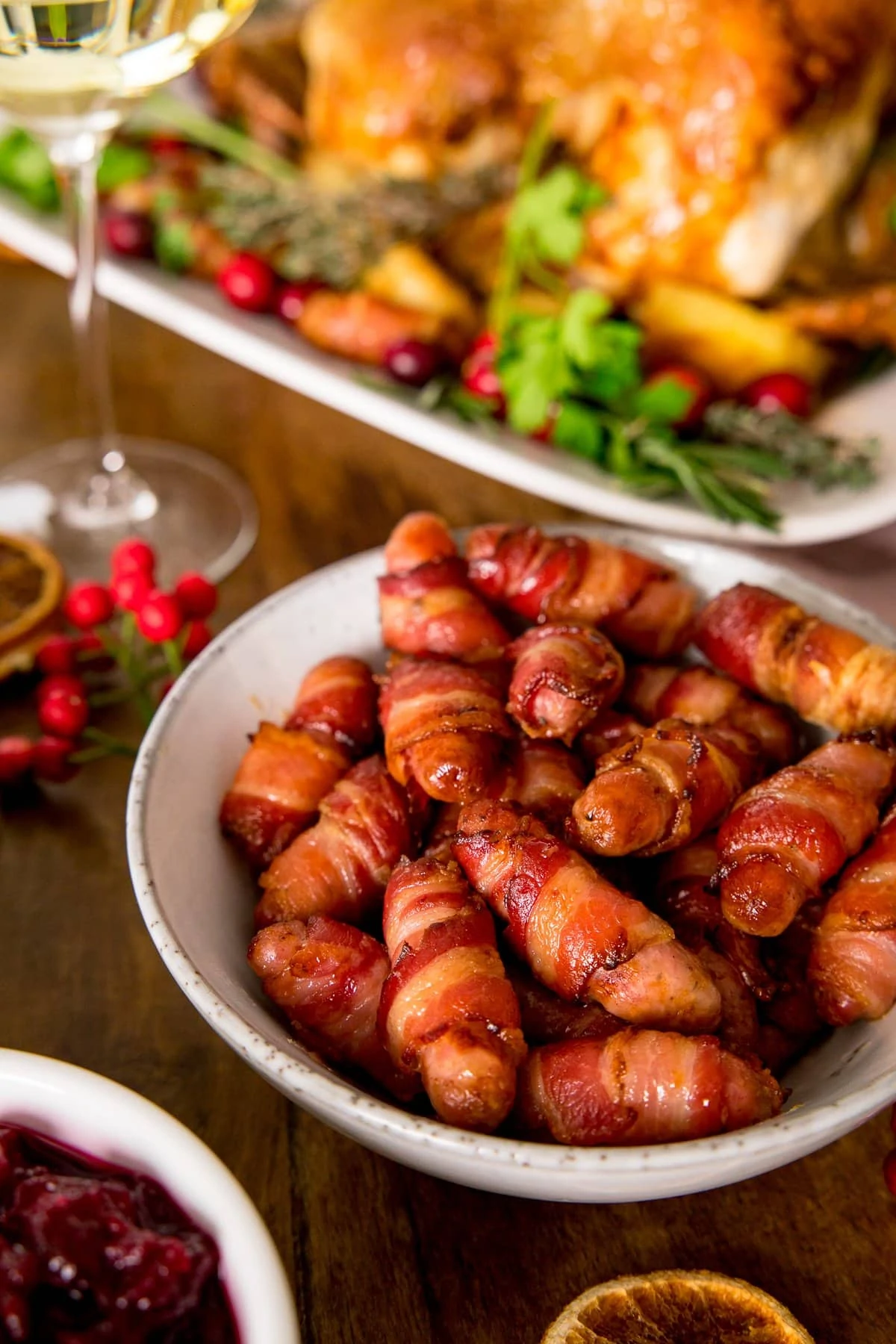 Cocktail sausages wrapped in bacon (pigs in blankets) in a white bowl on a table with other Christmas dishes partially in shot