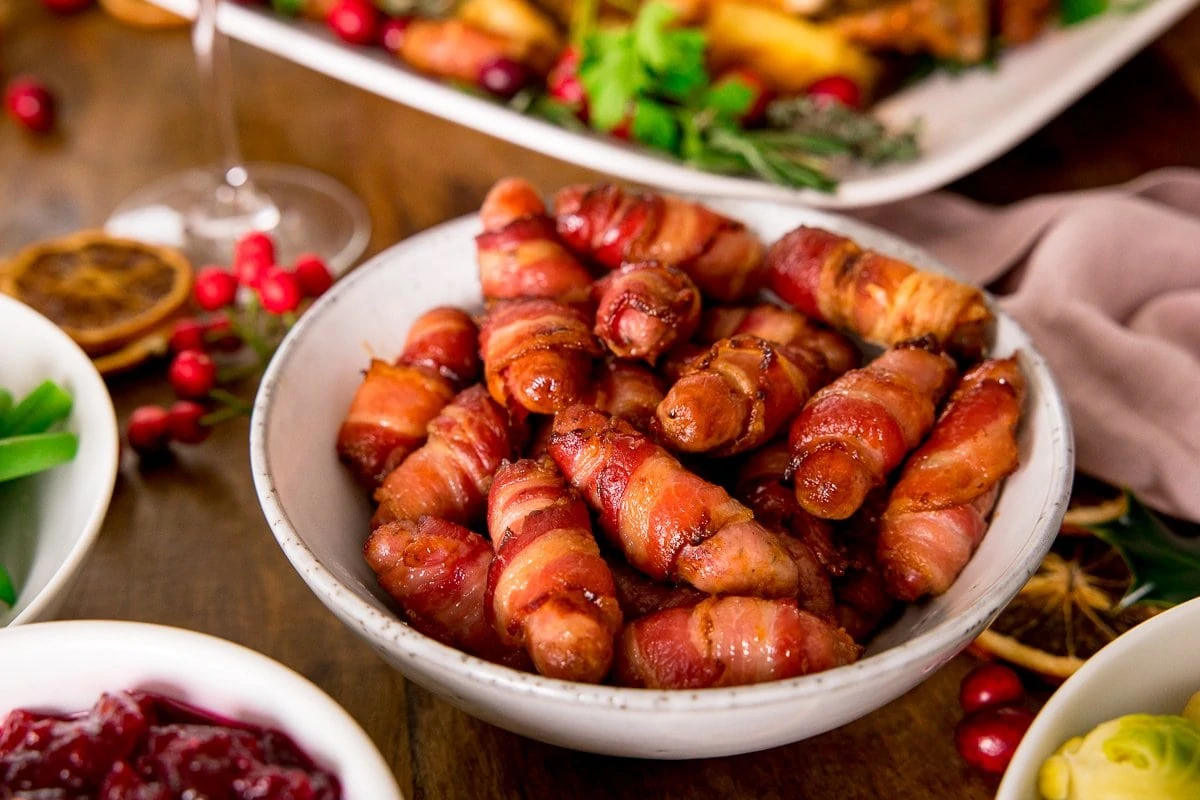 Bacon wrapped sausages in a white bowl on a table with other festive dishes partially in shot