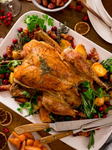 Roast turkey on a white plate surrounding by garnishings of herbs, cranberries and Christmas dinner side dishes