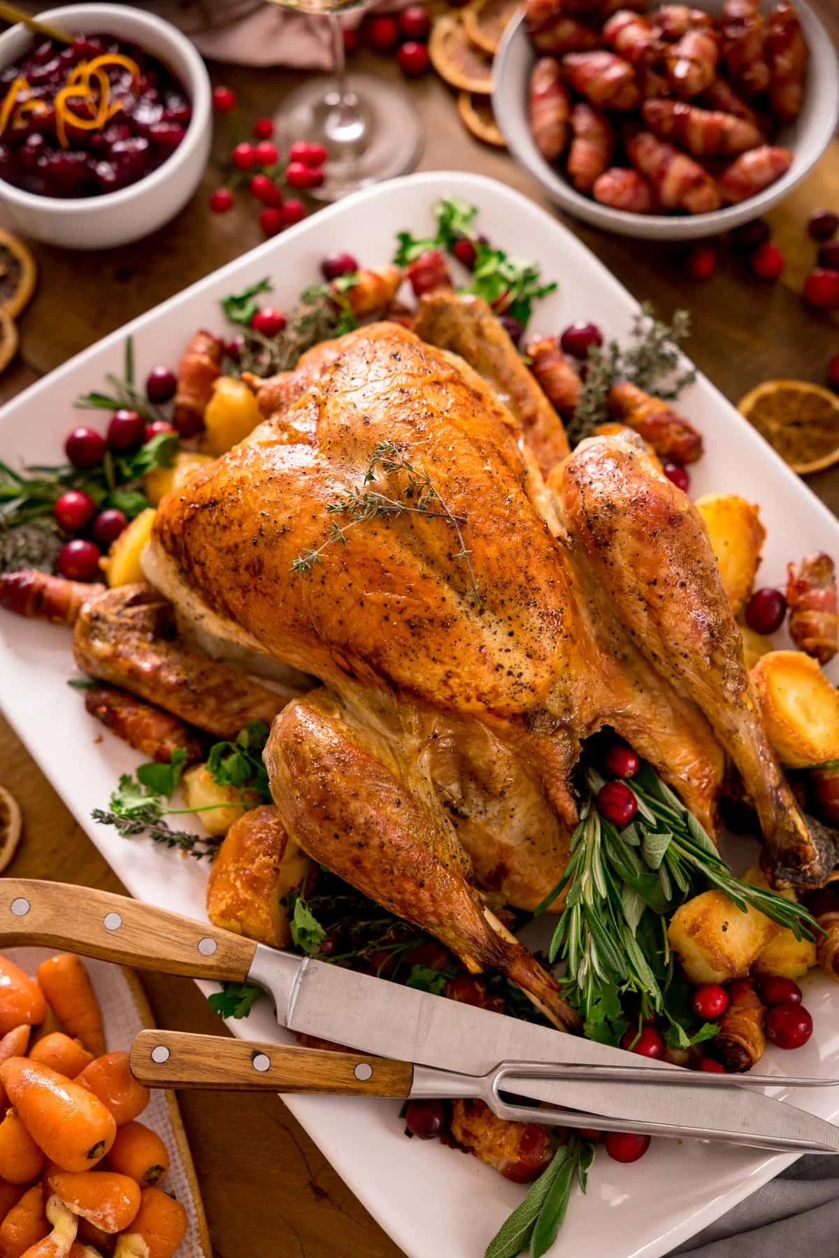 Roast turkey on a white plate with herbs, side dishes and a carving knife and fork on the plate.