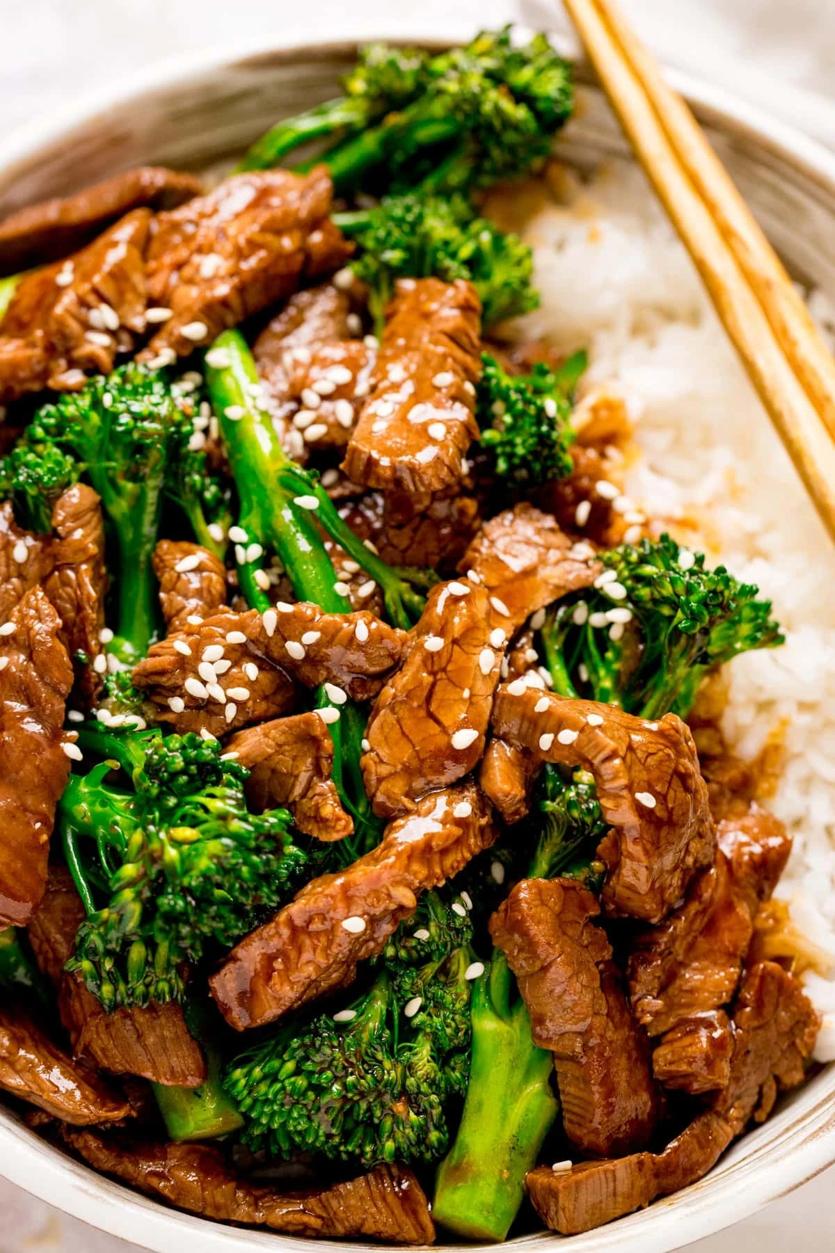 beef and broccoli topped with sesame seeds in a bowl with rice. Chopsticks resting on bowl.