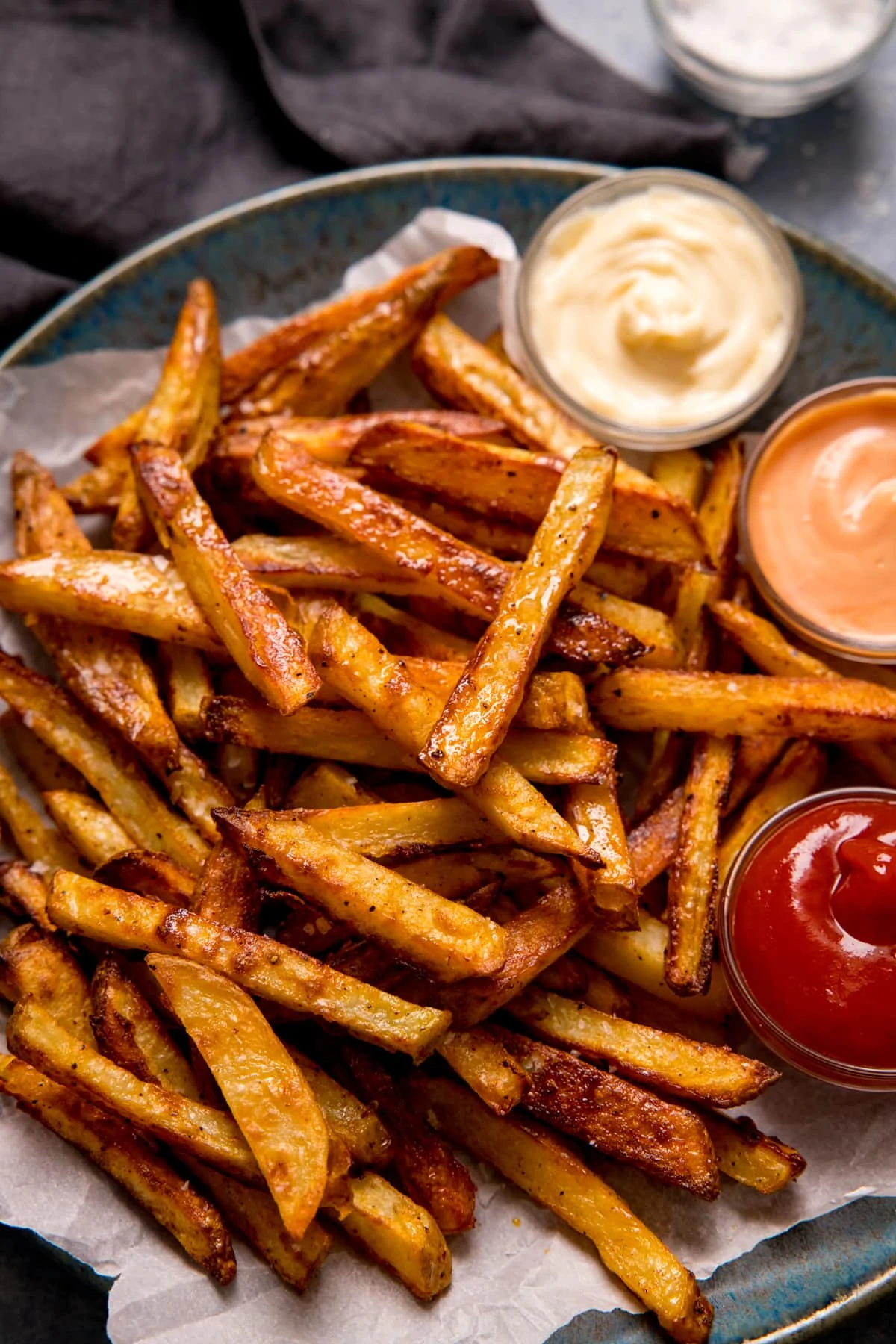 Crispy oven chips on parchment paper on a blue-grey plate. Three bowls of dips on the plate.