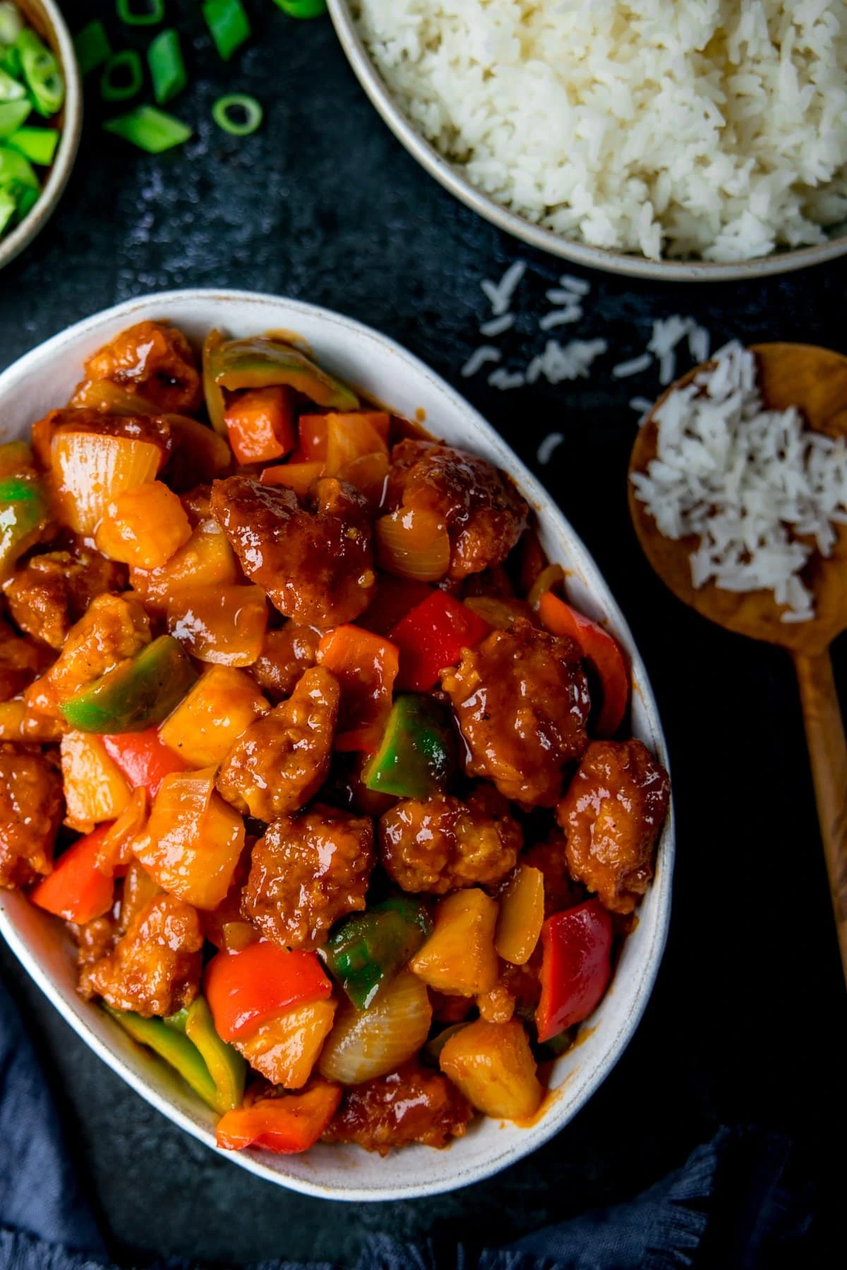 White oval bowl filled with sweet and sour pork. Rice and spring onions also on the table.