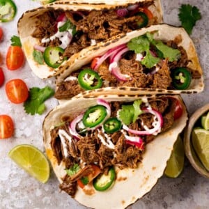 Square image of beef barbacoa on soft tacos topped with red onion and jalapeno slices